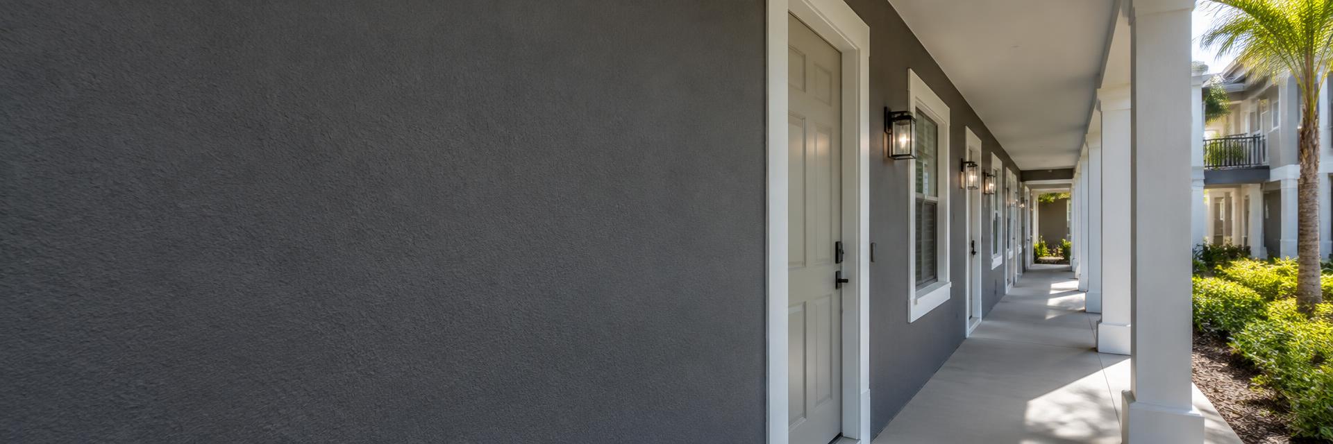 Multi-family apartment breezeway with gray painted stucco, white entry doors and columns overlooking a landscaped courtyard.